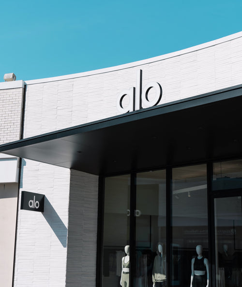 A photo of the front of the Alo Yoga Old Orchard store from a right angle showcasing the matte black awning and white stone façade with floor-to-ceiling front slider doors.  