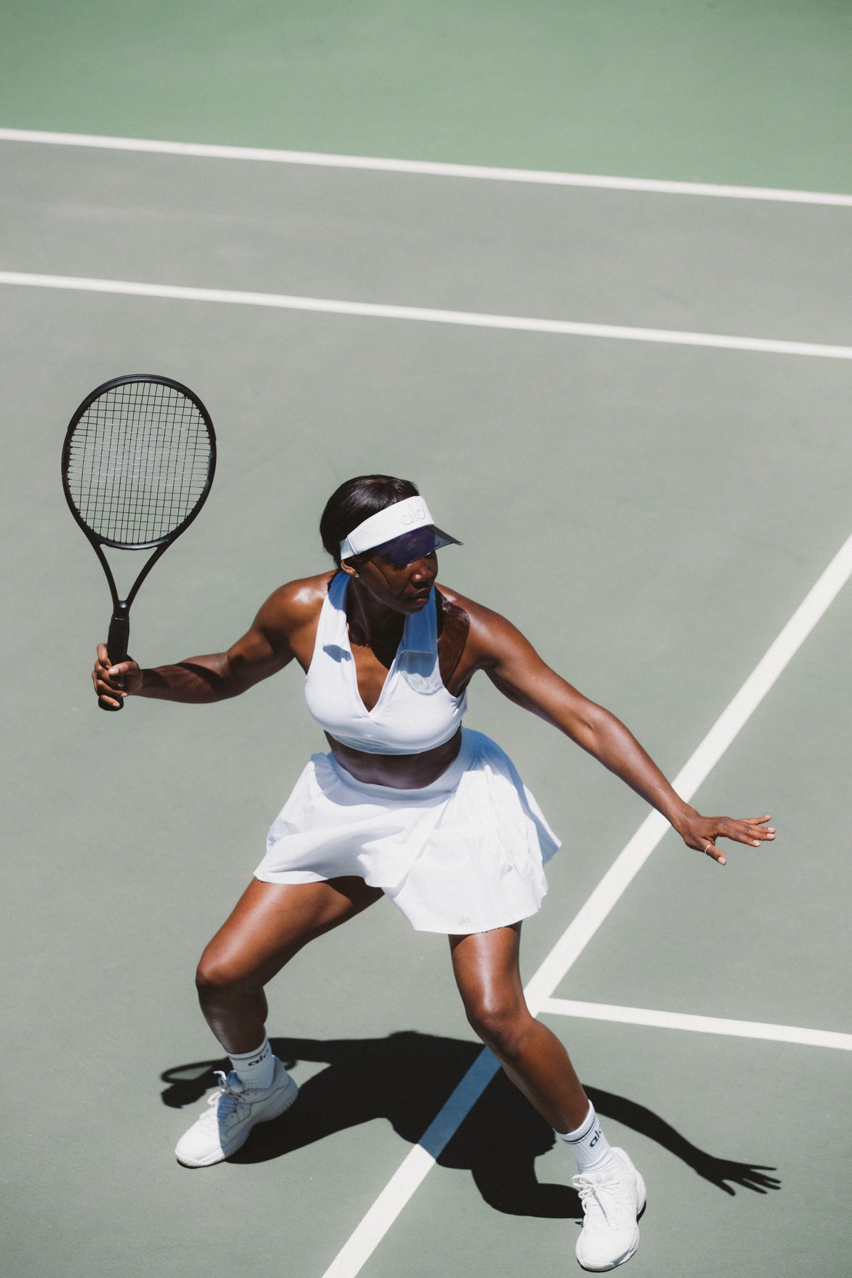 A woman wearing a white tennis skirt, a white collared sports bra, and a white and black visor while playing tennis. 