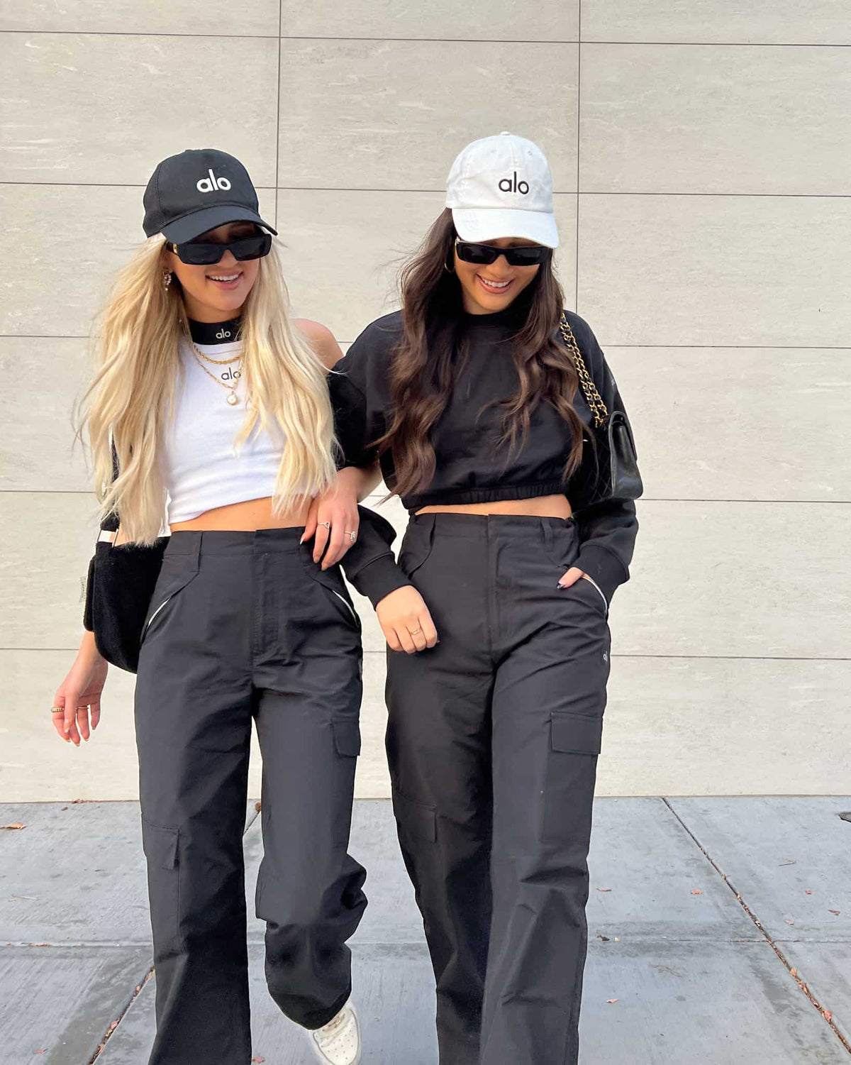 @fashionablysurfed and @shestyledwhat both wearing a pair of Black Edge Trousers with Alo hats and cropped tops walking arm in arm down a street sidewalk. 