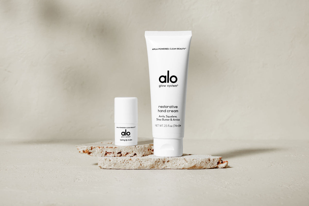 A photo of Alo’s Restorative Hand Cream and Lasting Lip Balm positioned on a tan stone.  