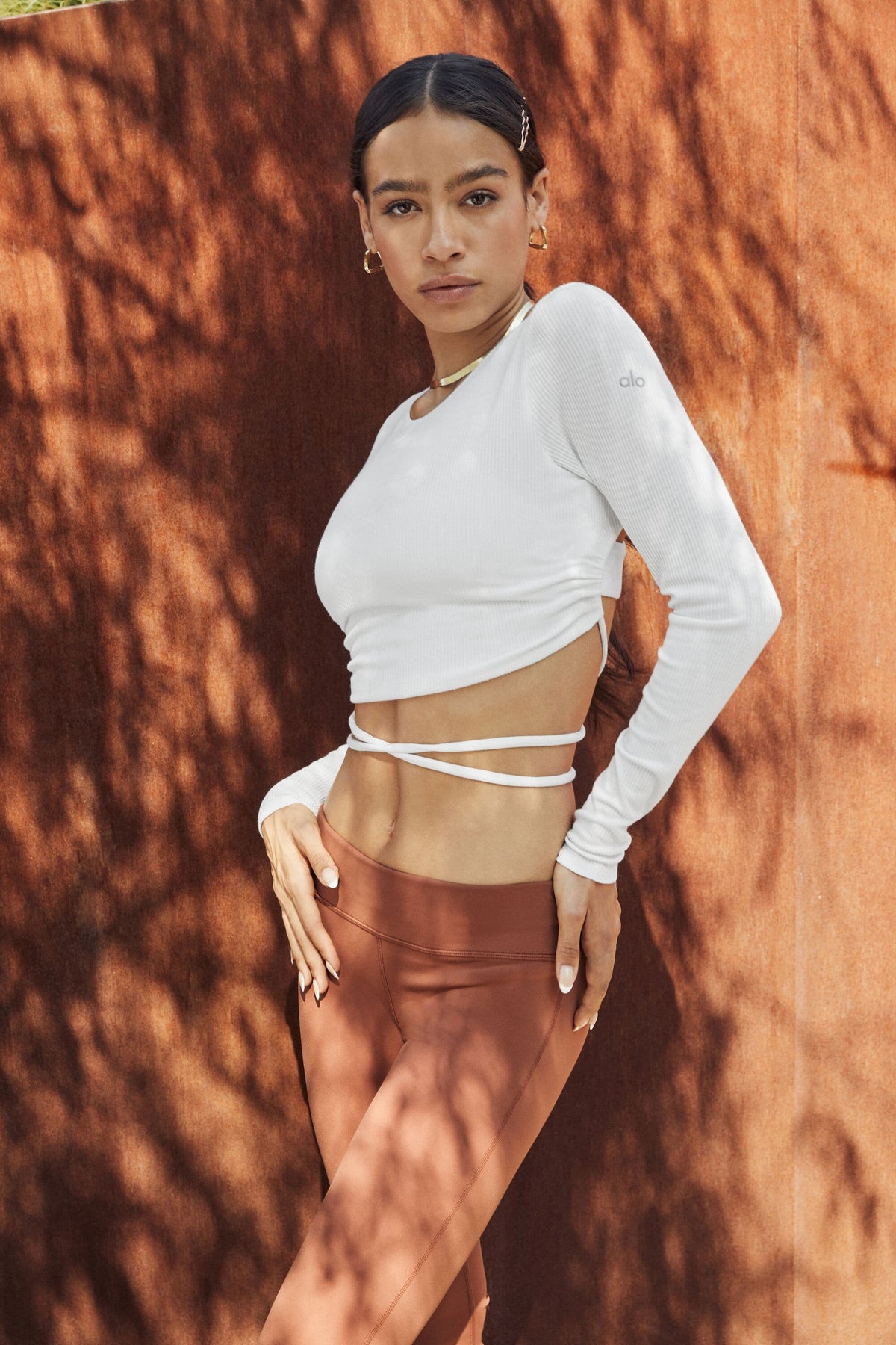 A woman wearing a pair of Rust Airbrush Low Rise Leggings with a White Ribbed Manifest Long Sleeve Top while posing against a rust-colored wall.  
