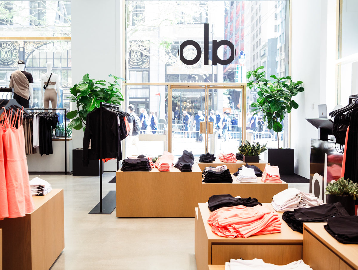 A photo taken inside of an Alo Yoga store facing the storefront with the Alo logo hung above the door, natural wood blocks displaying the apparel, and two large fiddleleaf trees framing the front entry. 