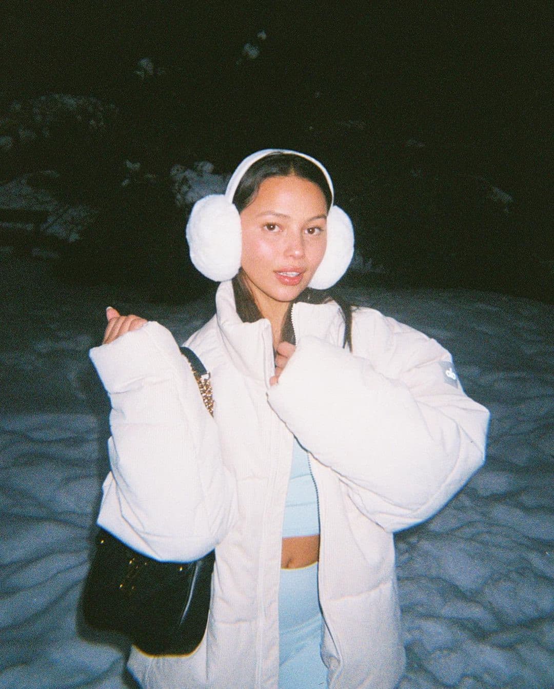 @fiona.bl wearing a light blue workout set with a sherpa puffer jacket on top and faux fur ear muffs while standing on a snow-covered ground at night.