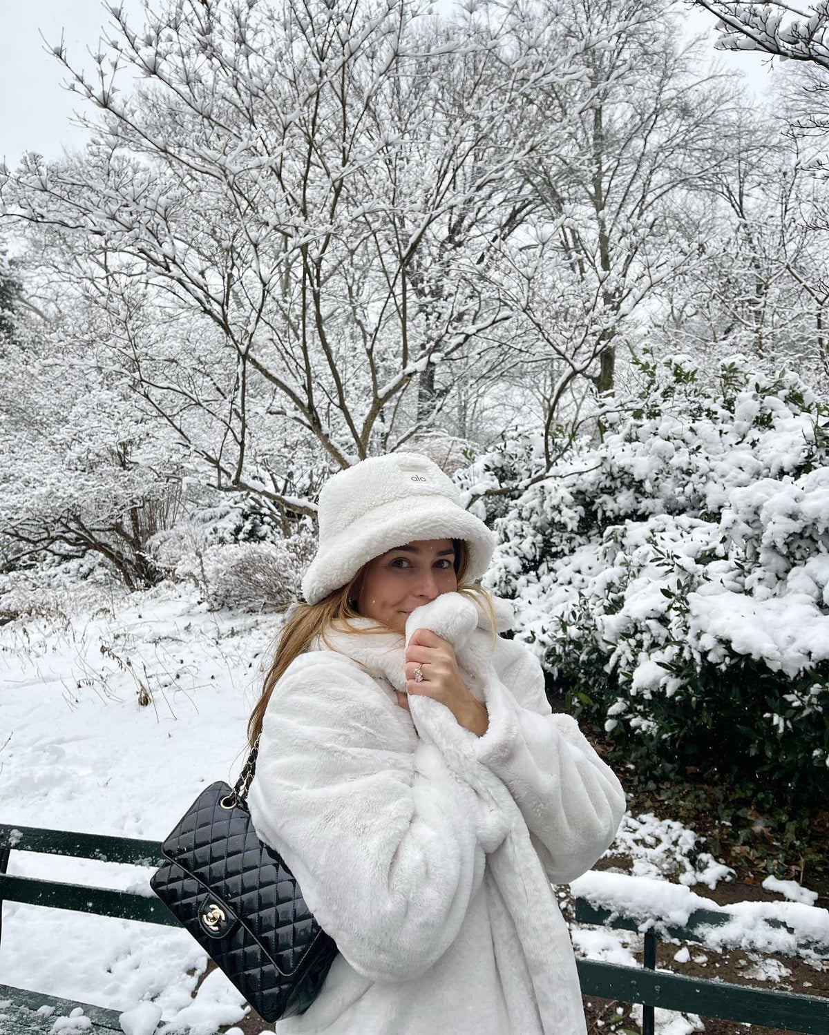 @coralsimanovich wearing a white faux fur trench coat with a sherpa bucket hat and a Chanel purse while walking a snowy park.  