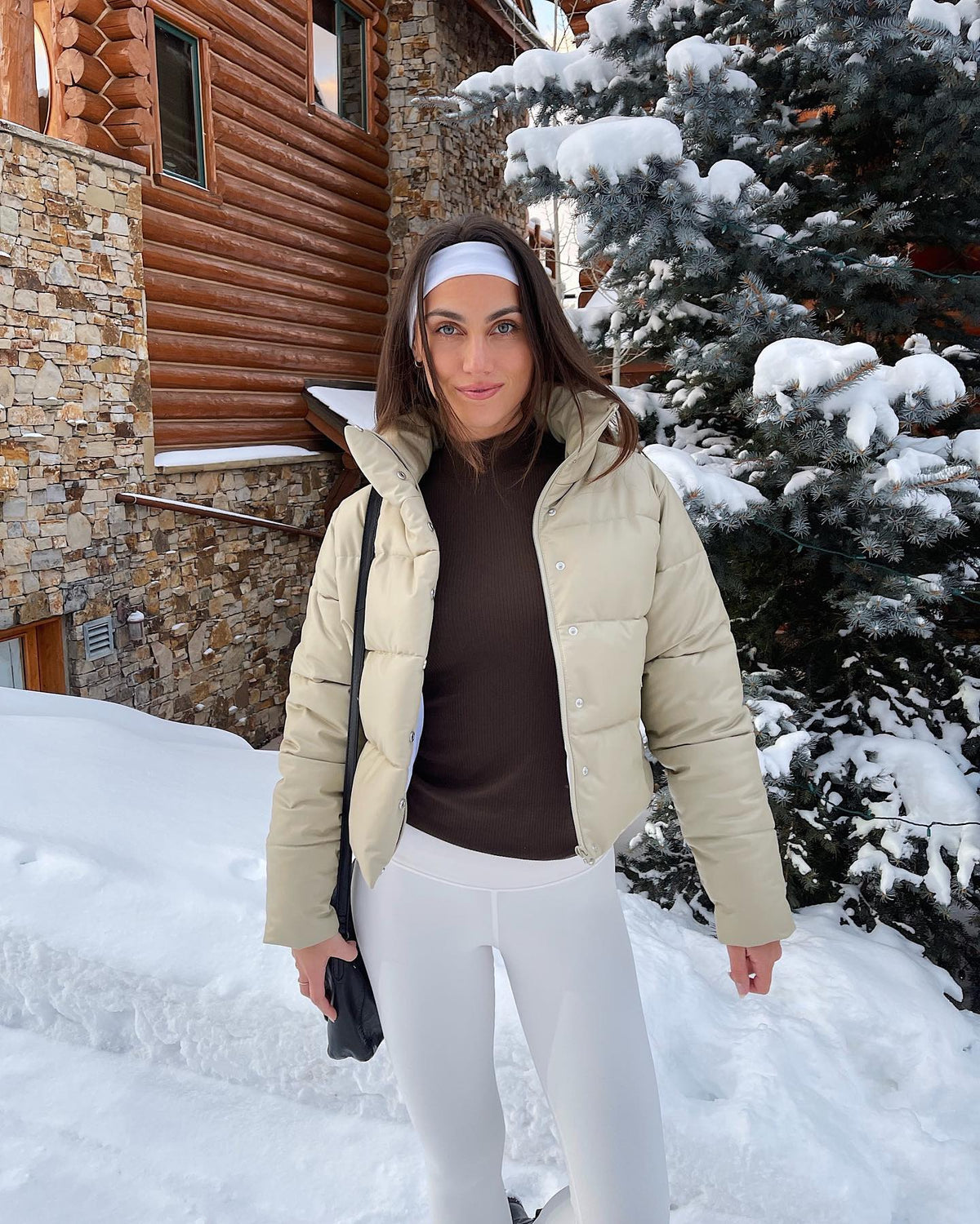 @stephjrayner wearing a Faux Leather Gold Rush Puffer in California Sand paired with a pair of High Waist Airbrush Leggings while standing in front of a wood cabin and snow-covered pine tree.  