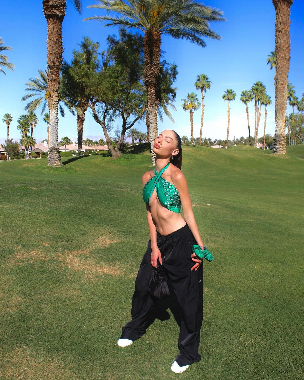 @iyardenedri wearing a pair of Courtside Tearaway Snap Pants with an Emerald Green Bandana Scarf and Scrunchie while posing on a golf green before heading to a music festival. 