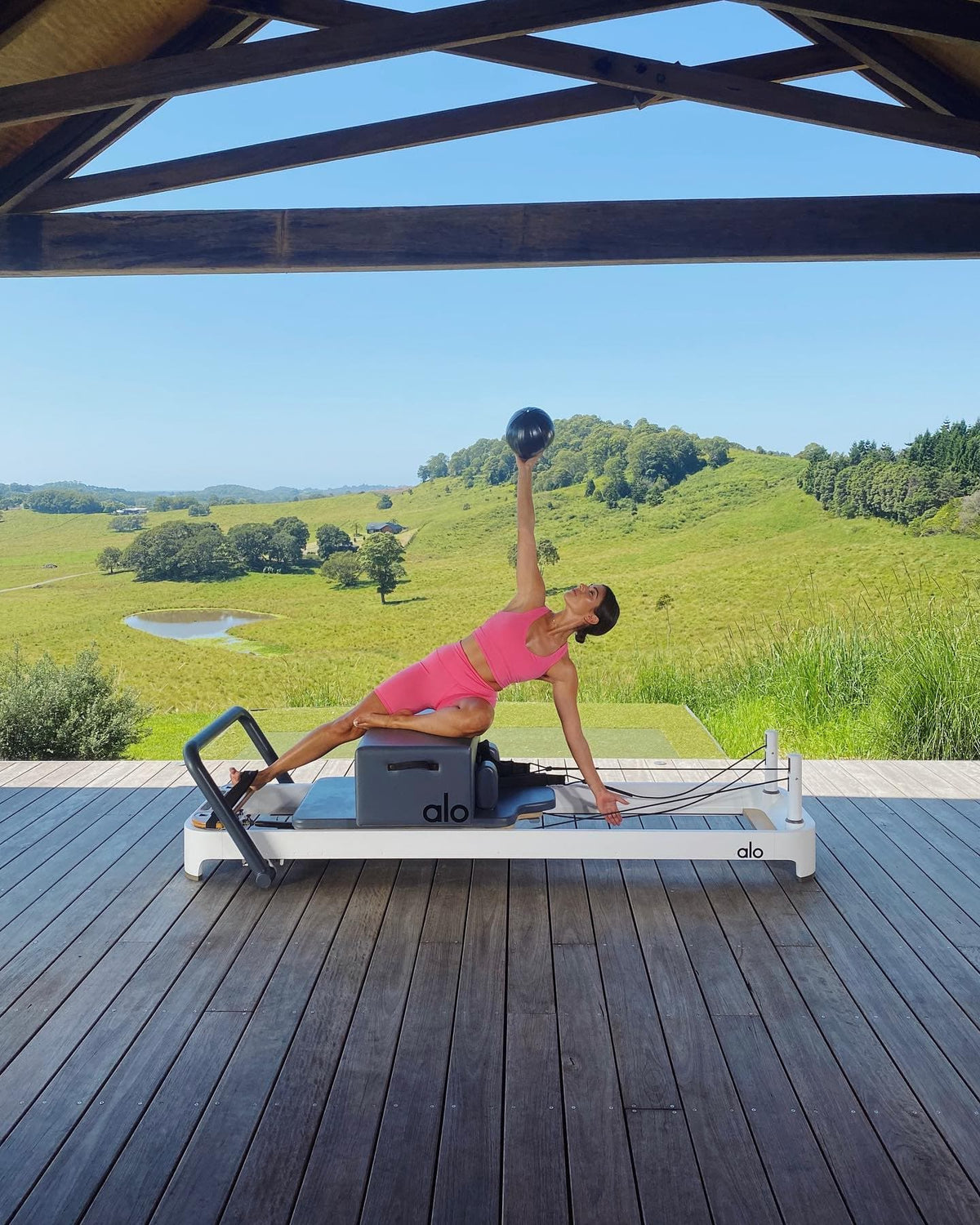 @biancamelas wearing an Airbrush Real Bra Tank with a pair of matching High Waist Biker Shorts in Pink Fuchsia while doing Pilates on a reformer with an exercise ball in a vast prairie landscape.  