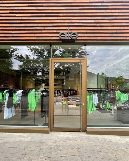 A photo of Alo Aspen’s storefront displaying wooden slats on the top, floor-to-ceiling windows beside the glass door, and an array of neon green apparel displayed in the windows.  