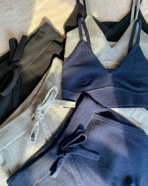 A flat lay photo of three cashmere bras and three cashmere pants in grey, black, and navy blue laying atop a white linen sheet. 