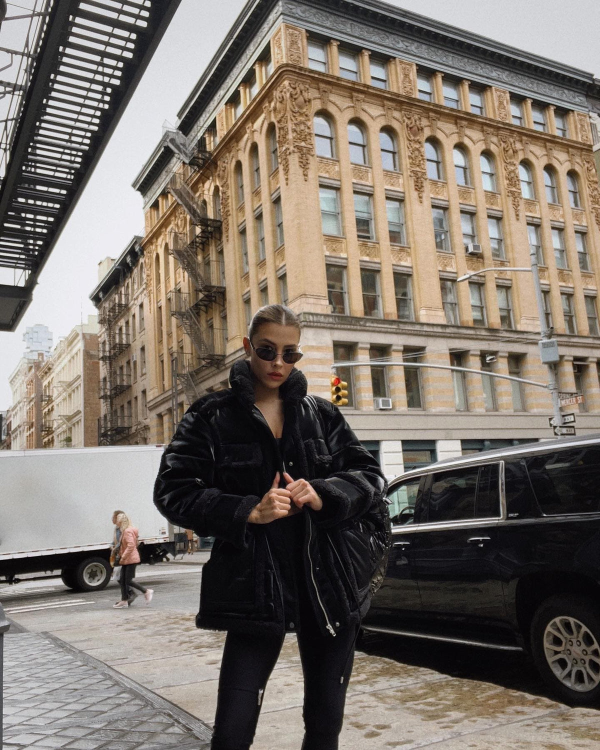 @michellesalasb wearing a black satin faux leather coat with sherpa details and a longer length paired with black leggings standing in front of a large building in the city. 