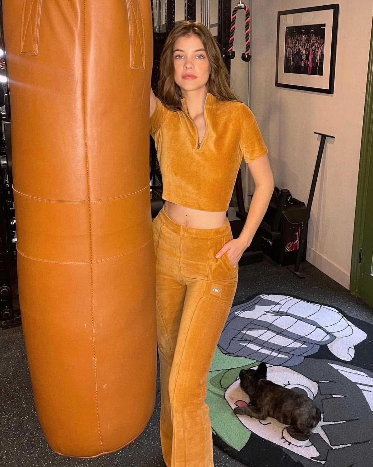 @realbarbarapalvin wearing a velour bootcut pant with a velour cropped quarter zip short sleeve shirt while posing next to a leather punching bag. 