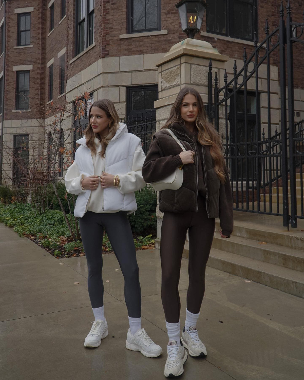  The twins of @babesontrend wearing matching ribbed puffer vests with a hoodie layered underneath and high-waisted leggings on the bottom while in front of a brick and stone building. 
