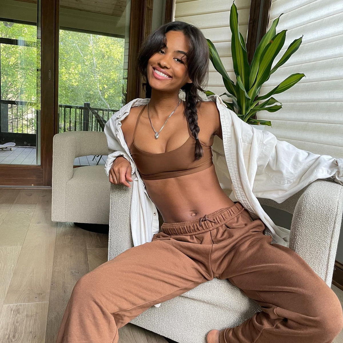 @_kendrabailey wearing a pair of brown straight leg sweatpants with a matching sports bra and a white oversized collared shirt on top while sitting on a boucle chair smiling for the camera. 