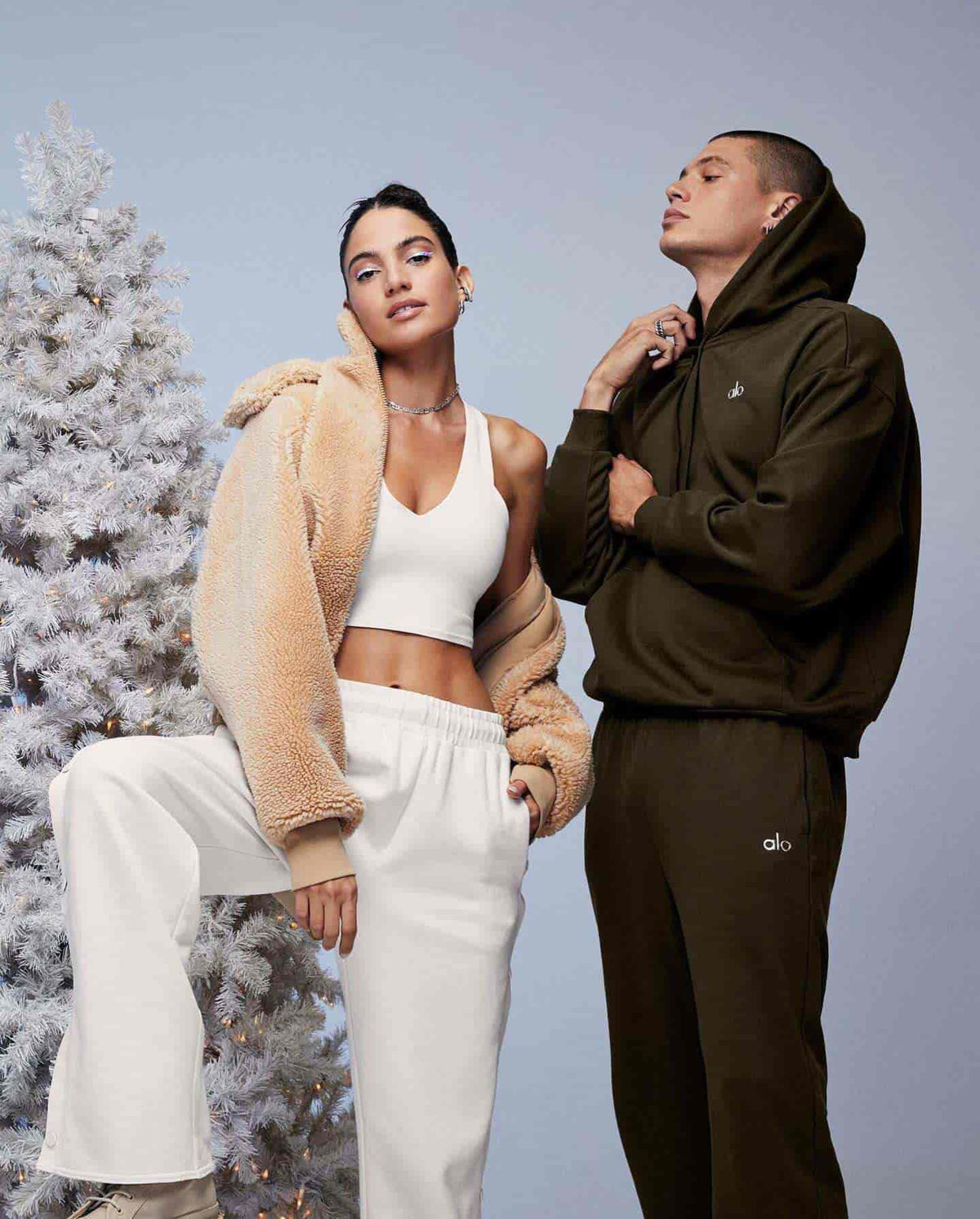 A man and a woman standing against a grey background and a frosted pine tree while bundled up in Alo Yoga's cozy loungewear posing for the camera.  