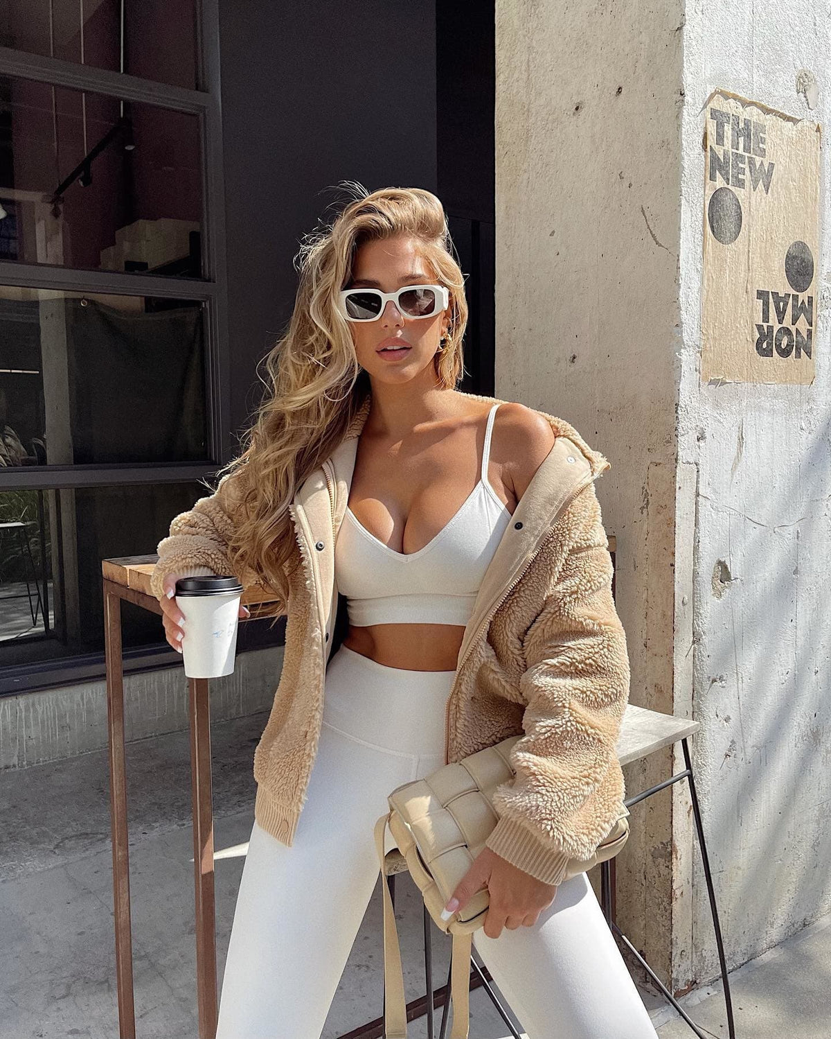 @karajewelll waring a tan sherpa jacket with snap buttons on top of a matching ivory workout set while holding a coffee and sitting on a barstool outside of the coffee shop. 