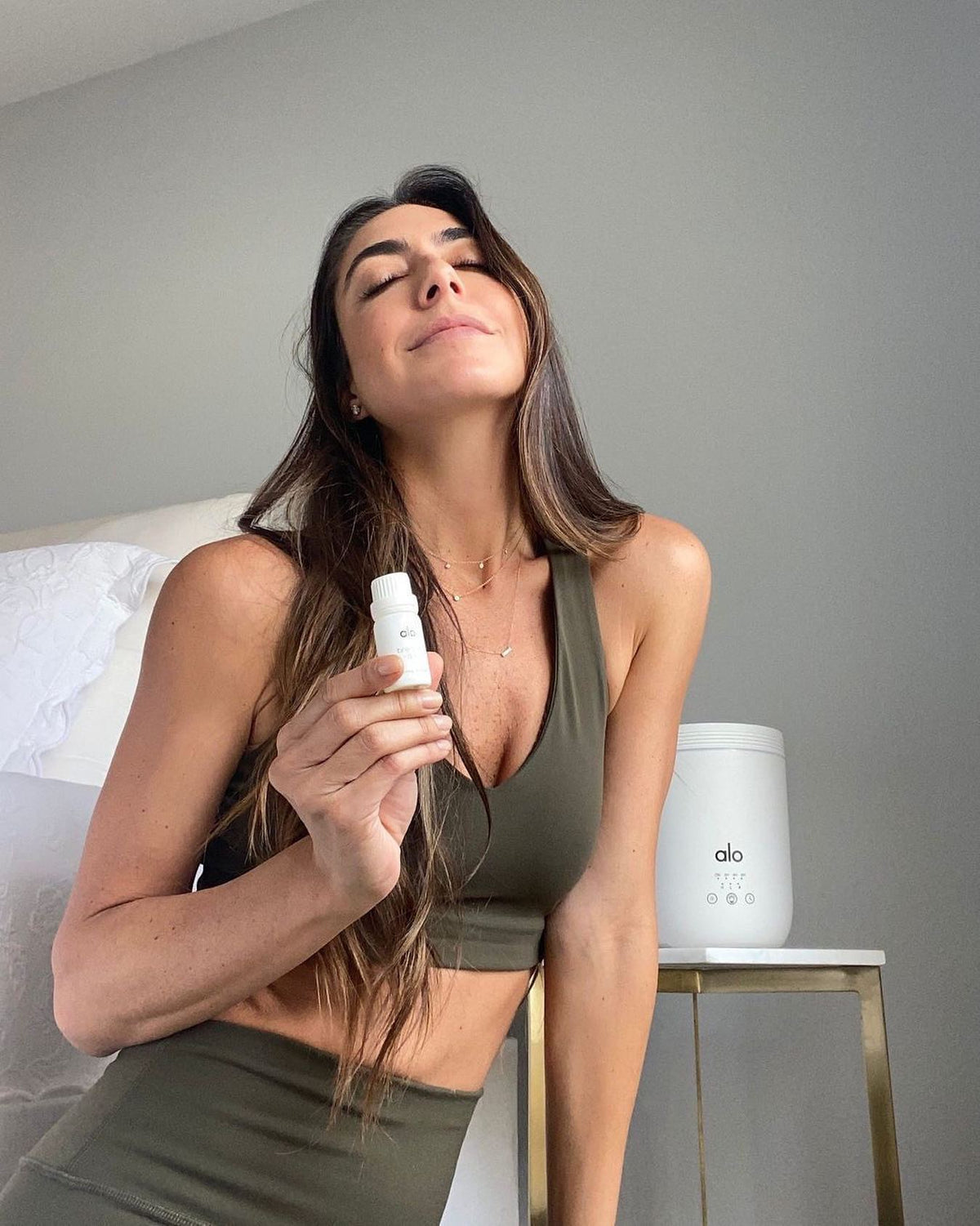 @revolveactive post of woman with long brown hair posing with Alo essential oil bottle and aura diffuser behind her on an end table. 